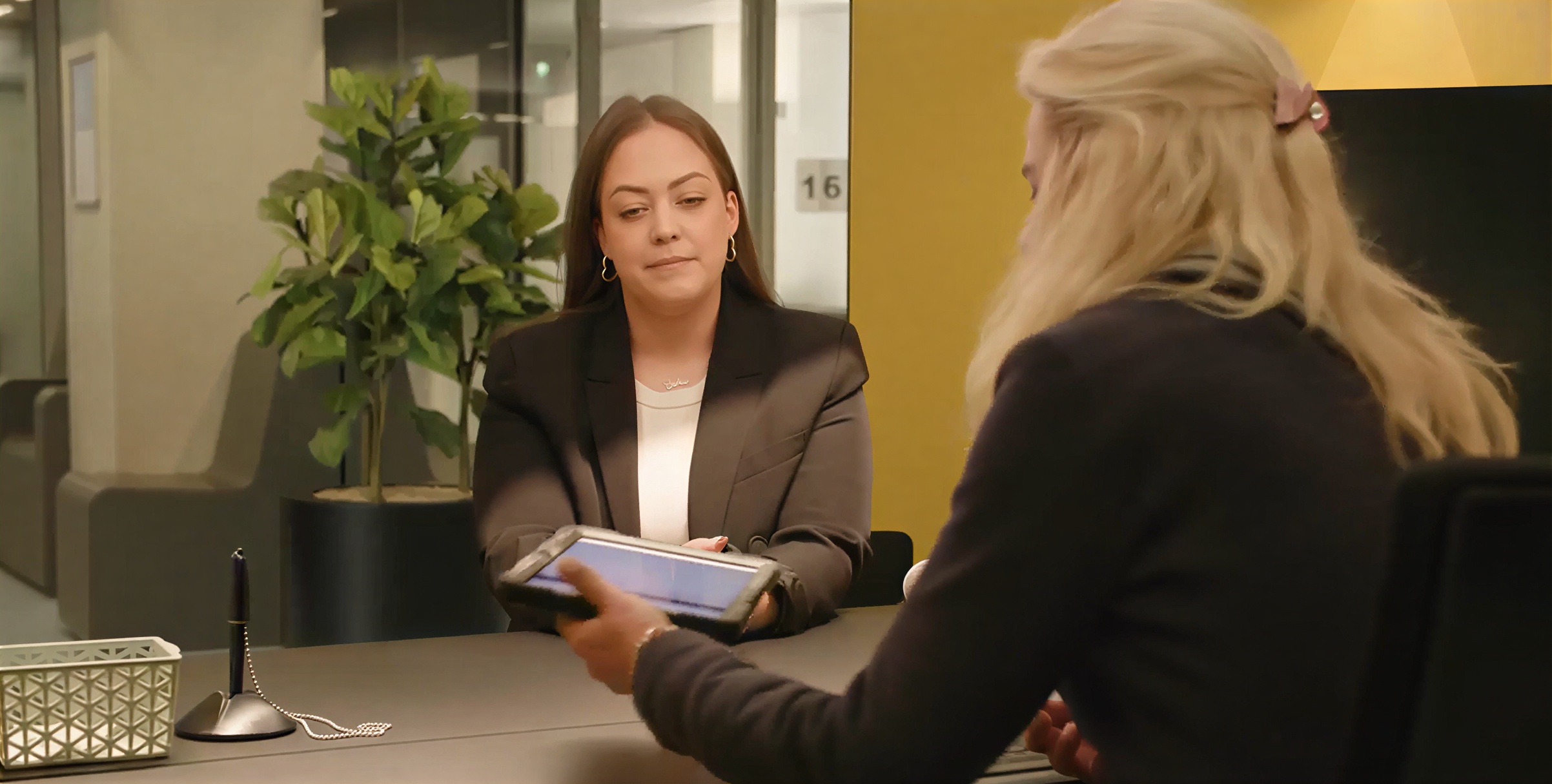 Video still: an IND employee seated at a desk hands a tablet in a protective case to a person opposite her, in an interview room at Ter Apel. The tablet screen is not legible.